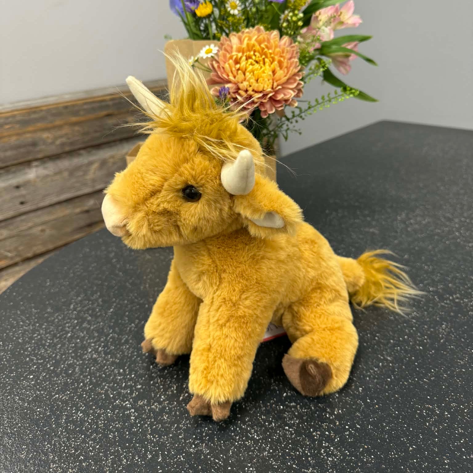 Plush highland cow toy on a dark surface with a bouquet of flowers in the background.