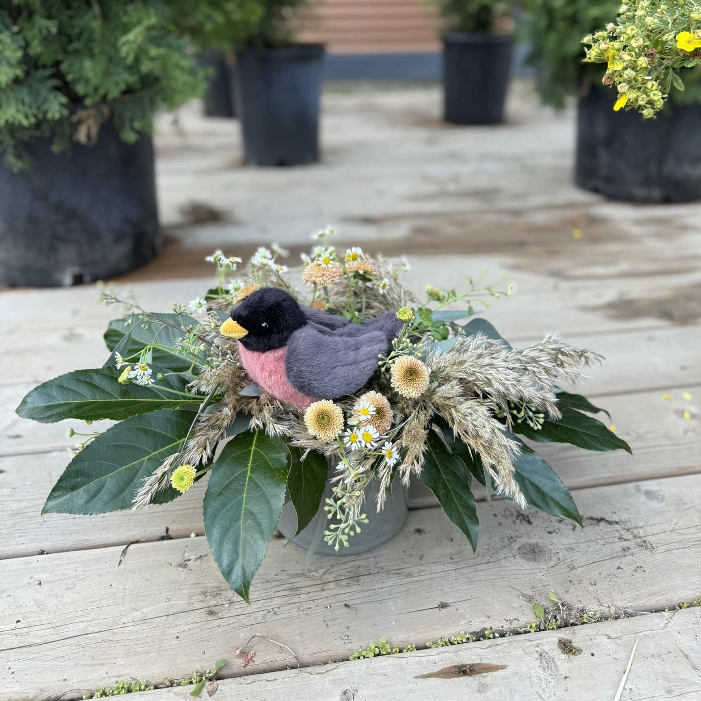 Decorative arrangement with a bird figurine and flowers on a wooden surface, with potted plants in the background.