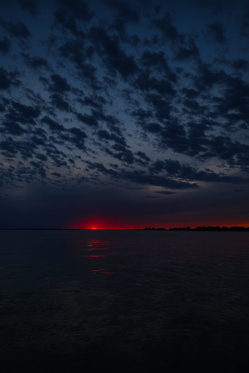 A scenic landscape photograph of a sunset over calm water, featuring a vibrant blue and purple sky filled with textured clouds, with a bright red and orange glow on the horizon reflecting on the water surface.