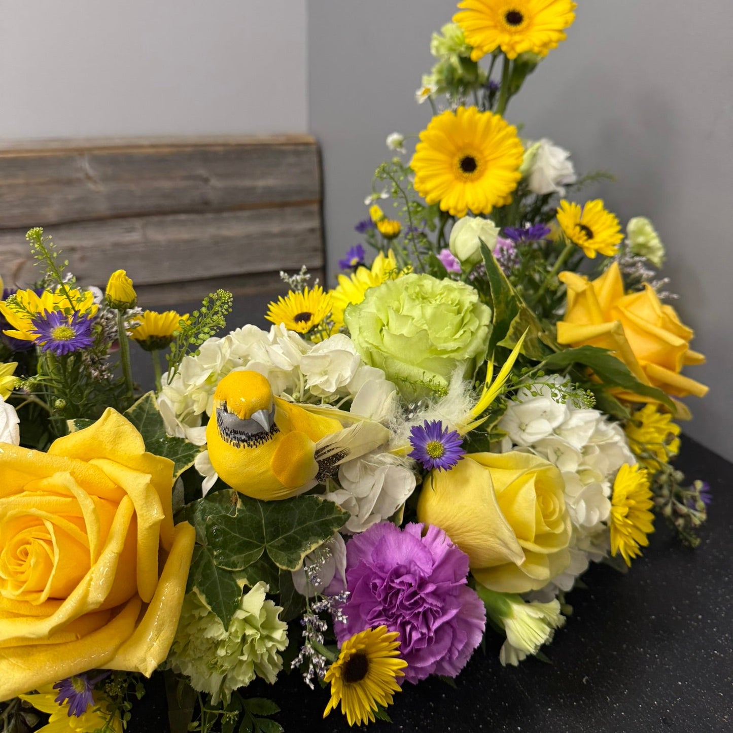 Floral arrangement with yellow flowers and a bird figurine on a dark surface.