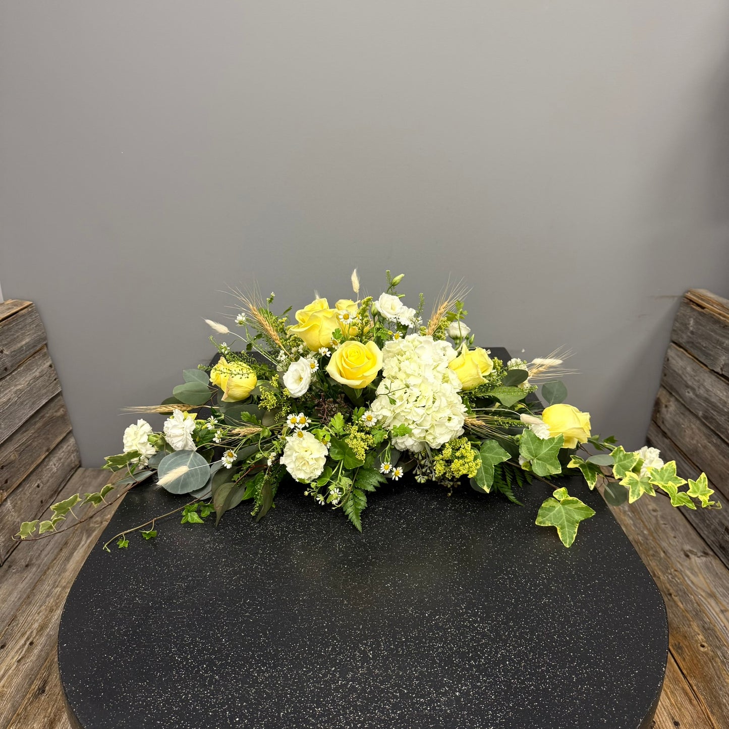 Floral arrangement with yellow and white flowers on a black surface against a gray wall.
