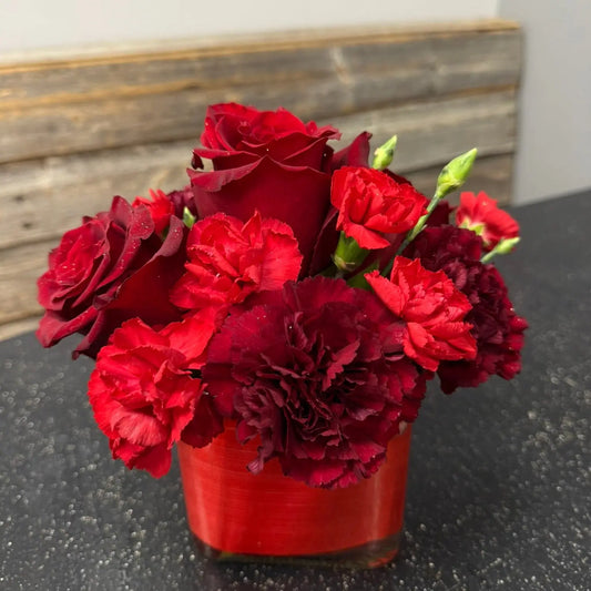 Bouquet of red and burgundy flowers in a red vase on a dark surface with a wooden shelf in the background.