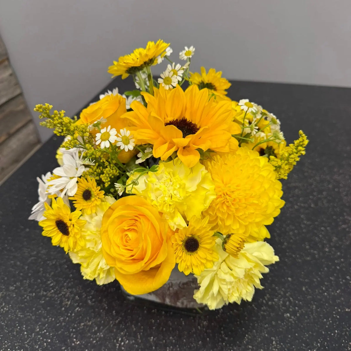 A vibrant yellow and orange flower bouquet featuring sunflowers, roses, and chrysanthemums in a clear glass vase, set on a dark speckled countertop against a neutral gray wall.