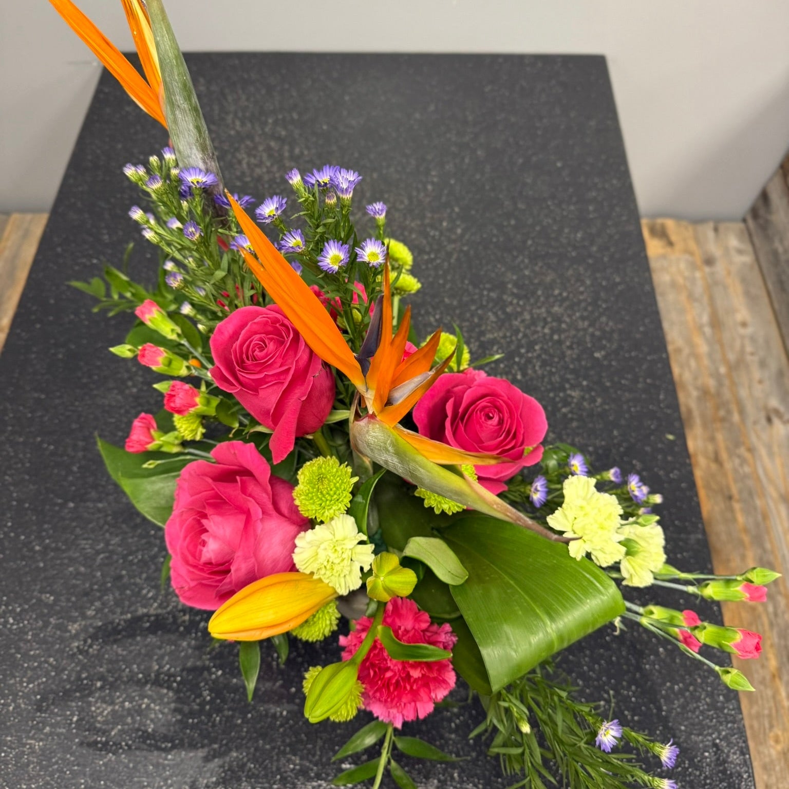 Colorful floral arrangement with pink roses, green leaves, and orange feathers on a dark surface.